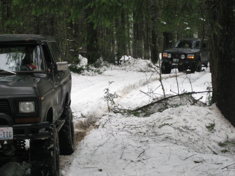 two trucks driving on a snowy forest road, one in foreground, one in background