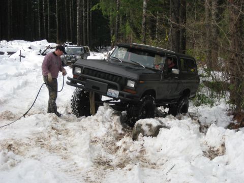 1979 Ford Bronco - A lifted off-road vehicle navigates a snowy, wooded trail with assistance from a tow rope.