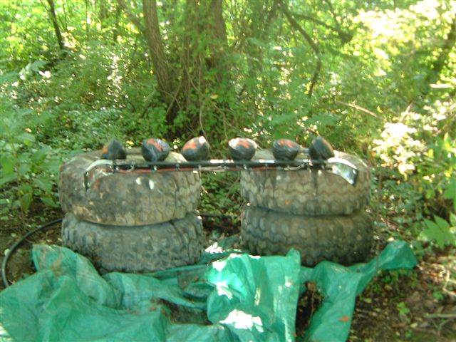Six off-road lights mounted on a metal bar above stacked tires in a wooded area