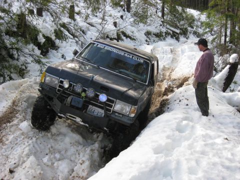 1979 Ford Bronco - A 4x4 navigates a challenging snowy path in a forest, with a bystander observing the off-road excursion.