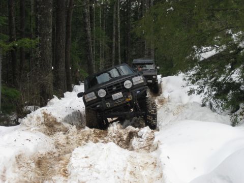 1979 Ford Bronco - Two modified trucks navigate a challenging snowy path through a dense forest.