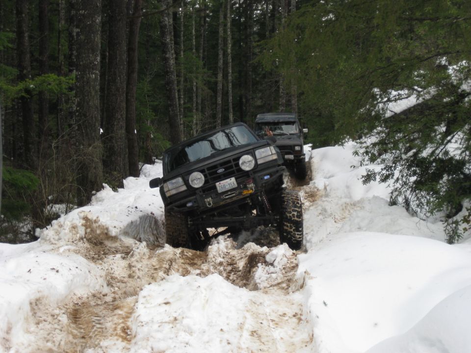 Two black off-road trucks driving through deep snow on a wooded trail. The front truck is angled, showing its lifted suspension and large tires