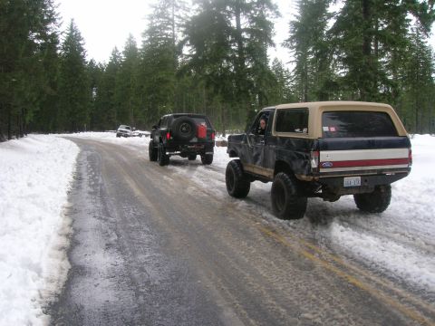 1996 Ford Bronco - Two SUVs drive on a wet and snowy road through a forest.