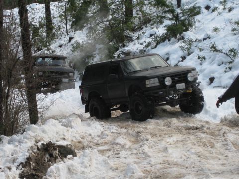 1979 Ford Bronco - Two off-road vehicles navigate a snowy trail, kicking up slush and snow.