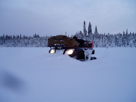 2000 Ford E350 - A four-wheeled vehicle is stuck in deep snow with its headlights illuminated, surrounded by a snowy landscape and a forest.