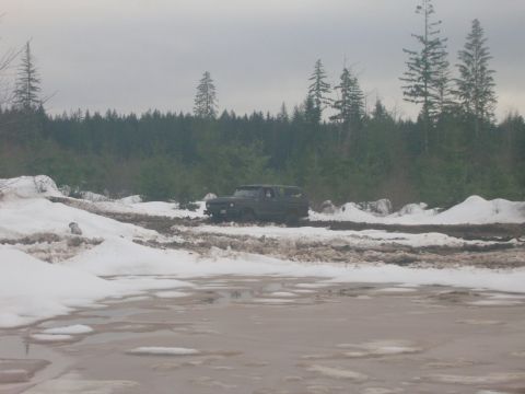 1996 Ford Bronco - A dark SUV navigates a muddy and snowy terrain with a dense forest behind it.