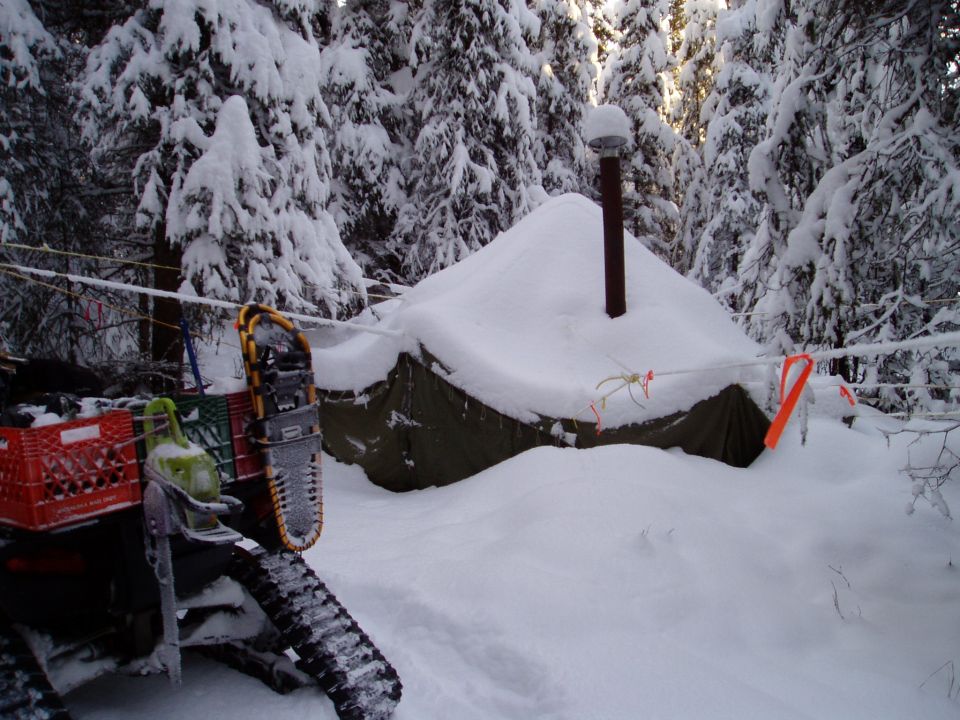 Snowmobile with red milk crates and snowshoes parked next to a snow-covered tent in a forest