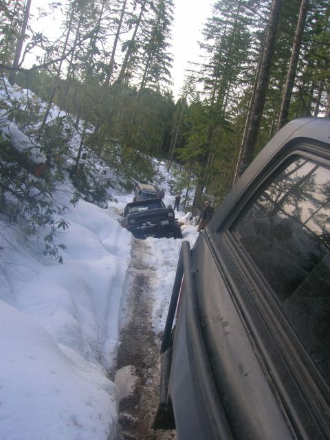 1996 Ford Bronco - Multiple SUVs navigate a challenging snowy path through a dense forest during an off-road excursion.