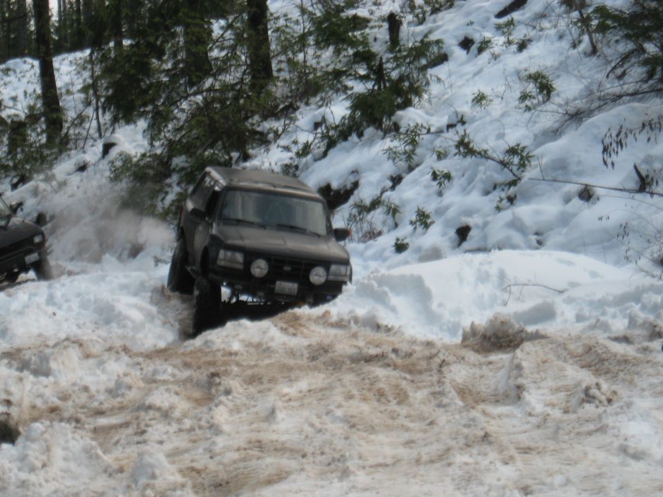 Dark SUV with off-road tires driving through deep snow, kicking up spray. Another vehicle is partially visible to the left