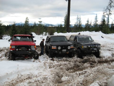 1989 Ford Bronco - Three trucks are stuck in a muddy, snowy area with trees and mountains in the background.
