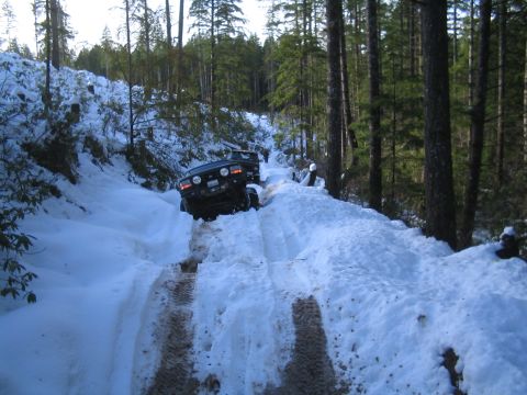 1989 Ford Bronco - Two SUVs navigate a challenging, snow-covered forest road, leaving tire tracks in the deep snow.