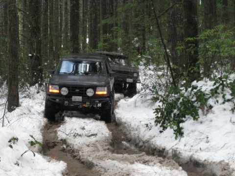 1979 Ford Bronco - Two Ford Broncos navigate a snow-covered forest trail, leaving deep tire tracks in the mud.
