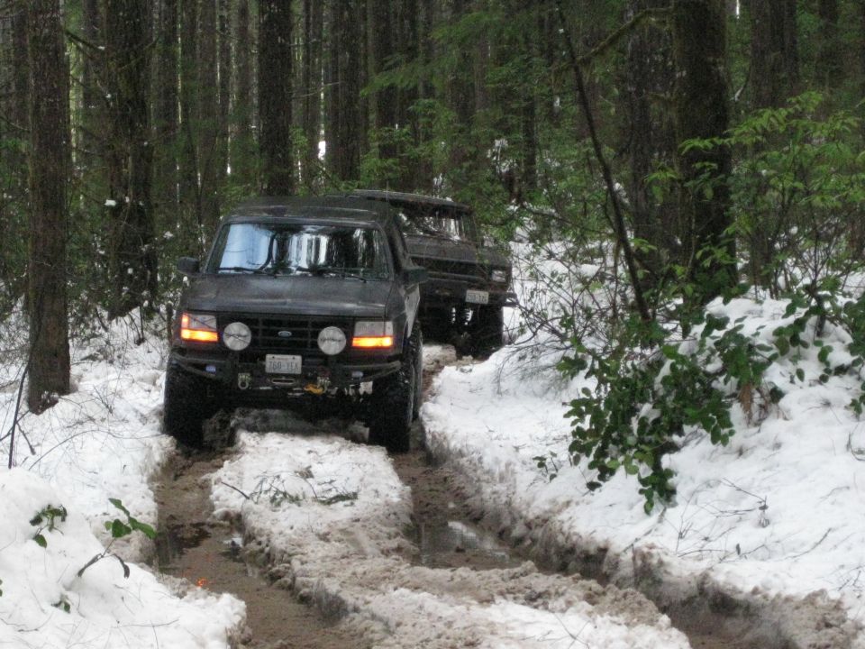 Black Ford Bronco driving on a snowy, muddy trail in a forest. Another Bronco is behind it