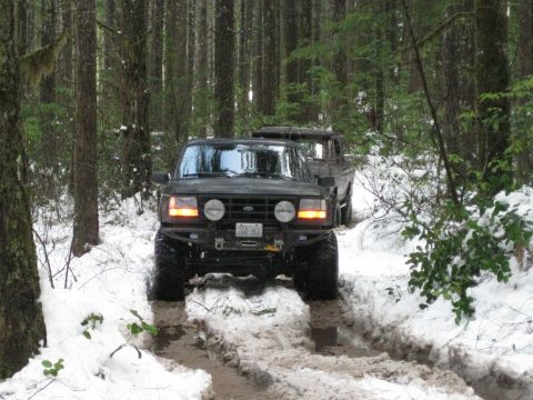 1979 Ford Bronco - A dark SUV navigates a snowy forest road, its lifted suspension and off-road tires evident as it drives through the snow.
