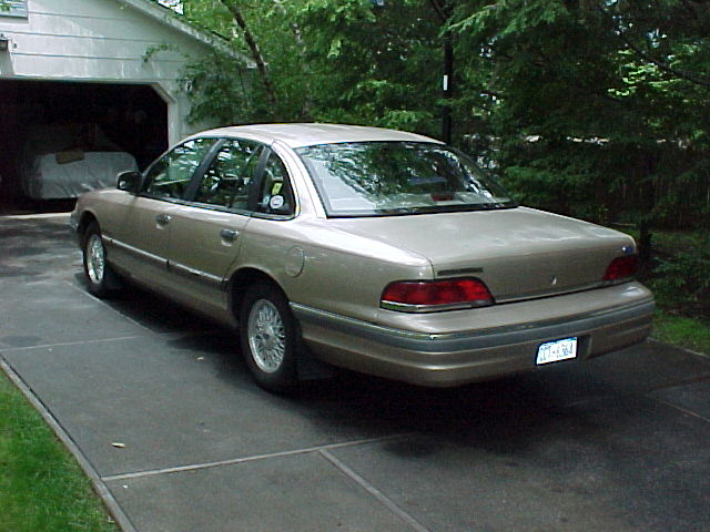 Gold Ford Crown Victoria sedan parked in a driveway, rear three-quarter view by a garage