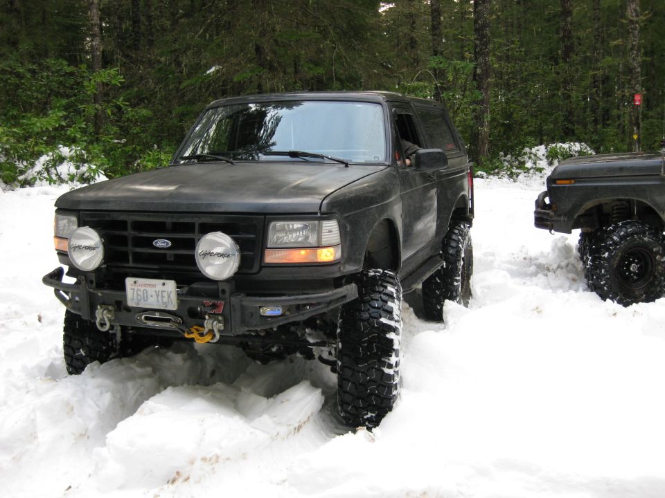 Black SUV with large off-road tires driving through snow, another vehicle visible to the right
