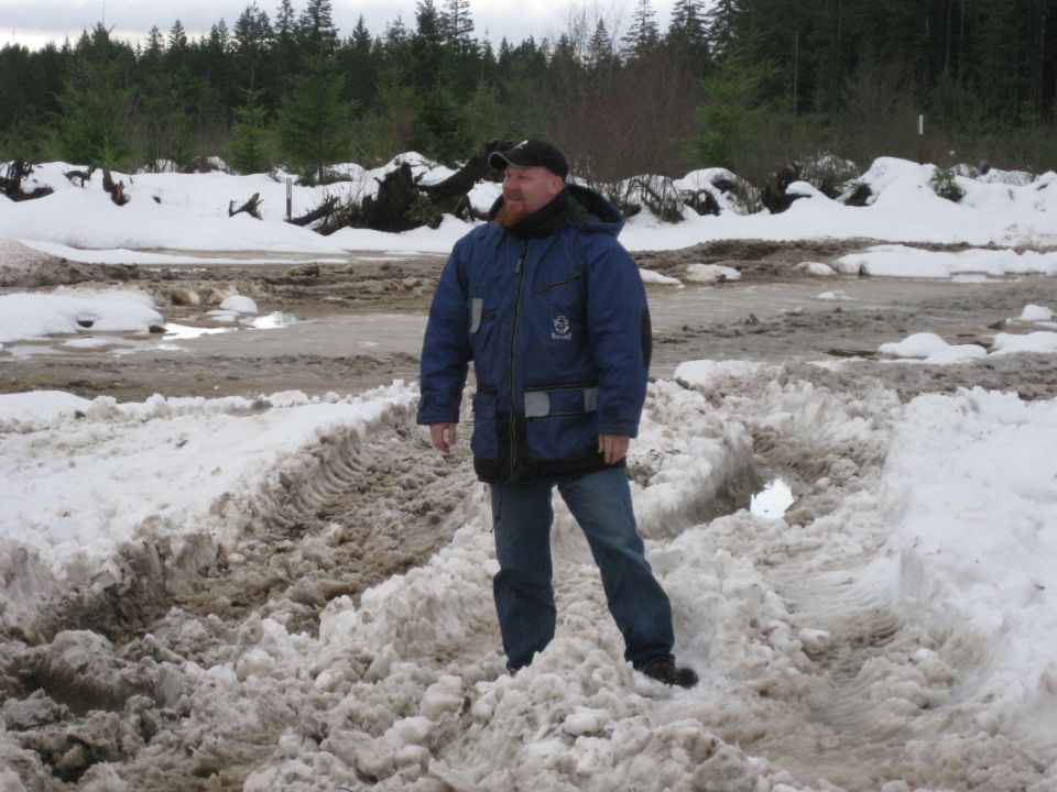 Man in blue winter coat and jeans stands in a snowy, muddy area with trees in the background