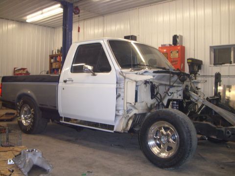 1994 Ford F-150 - A white regular-cab Ford pickup mid-teardown in a metal shop, stripped of its front clip, hood and engine so the bare frame rails, radiator…