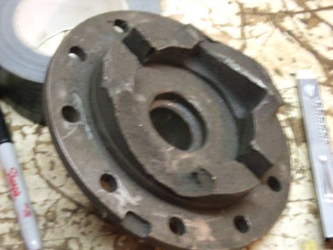 1996 Ford Bronco - A close-up, overhead view of a damaged, dark gray metal automotive part with several bolt holes around its perimeter and a central opening.…