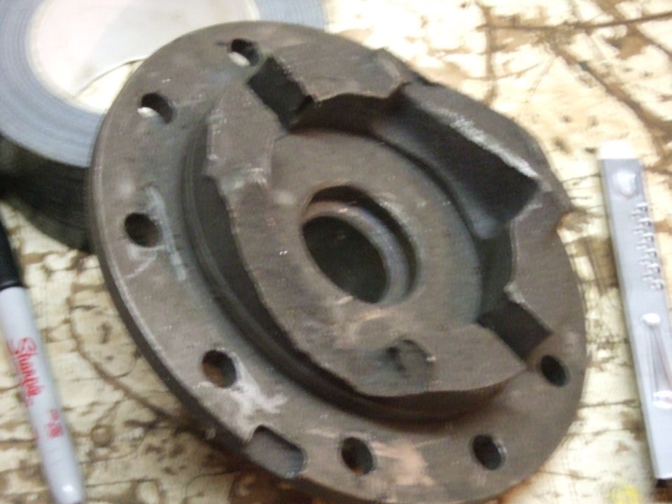 Damaged dark gray metal automotive part with bolt holes and central opening.