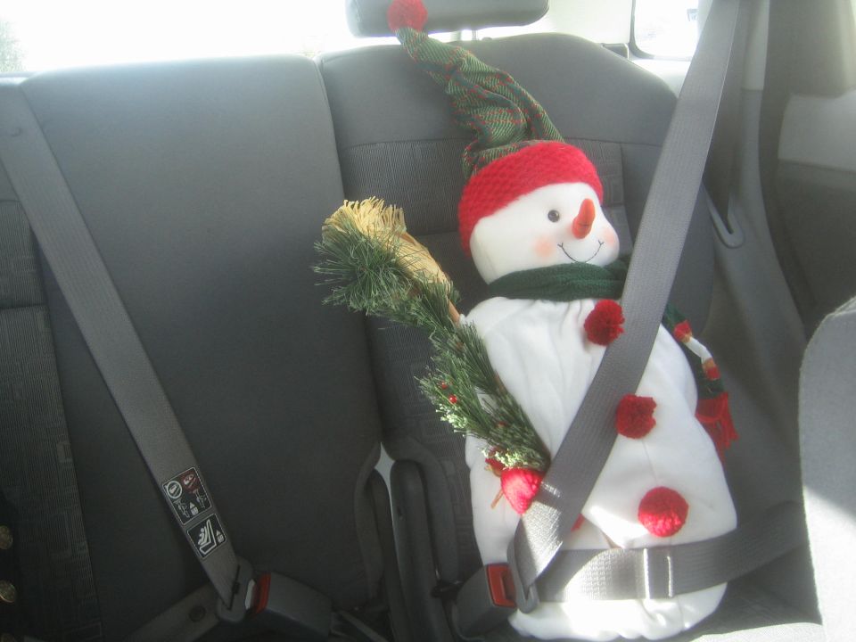 Snowman with a green scarf and red hat secured by a seatbelt in a car's back seat