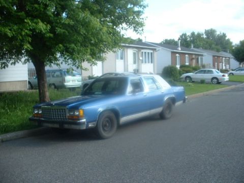 1987 Ford Crown Victoria LTD - A blue sedan is parked on the side of the road in front of houses.