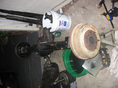 1989 Ford Bronco - A disassembled rear axle assembly with a brake drum and a bucket containing various parts sits on a garage floor.