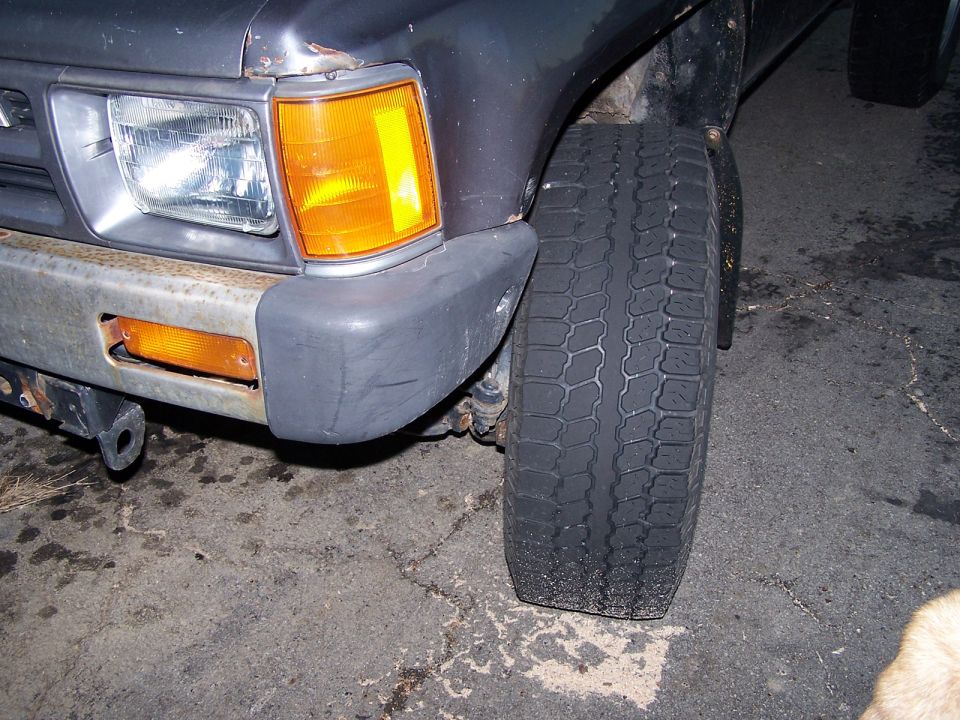 Front left corner of a gray Toyota pickup at night showing headlight, amber lights, rusty bumper, and front tire