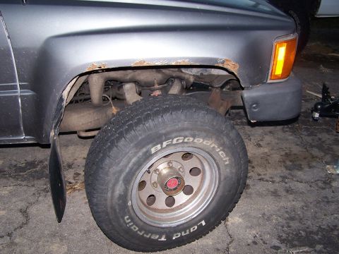 1988 Toyota Pickup/SR5 - Close-up of the front wheel well of a gray truck, revealing rust and a BFGoodrich tire.