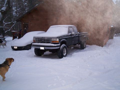 1986 Ford Bronco - A lifted Ford pickup truck and a car sit covered in snow outside a wooden building.
