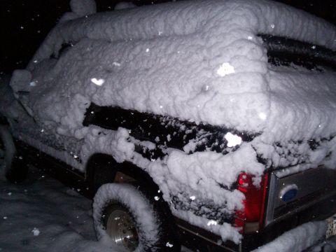 1986 Ford Bronco - A black 1986 Ford Bronco is heavily covered in snow, obscuring most of its details.
