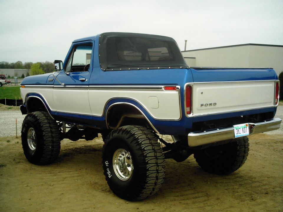 Lifted blue and white 1970s Ford Bronco with black soft top, rear three-quarter view