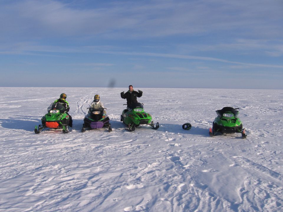 Four snowmobiles parked in a row on a frozen snowy expanse with two seated riders and one standing man