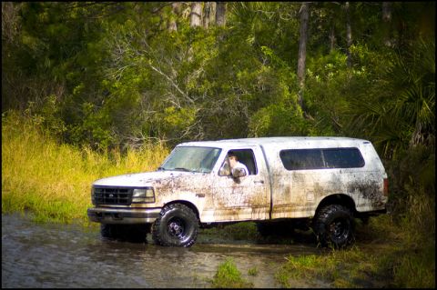 1996 Ford F-150 - A white pickup truck, covered in mud, drives through a shallow body of water with trees and vegetation in the background.