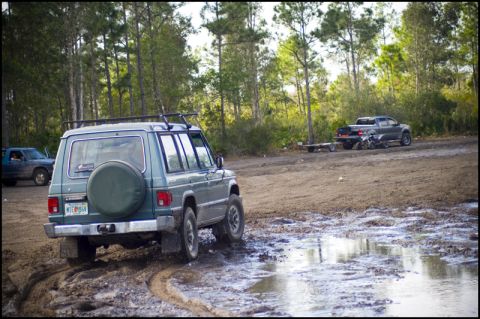 1996 Ford F-150 - An off-road scene with a Mitsubishi Montero navigating a muddy trail, while a pickup truck and trailer wait nearby.