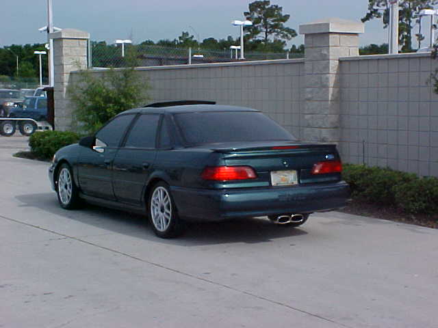 Dark green Ford Taurus sedan, rear three-quarter at dusk, dual exhaust and block wall behind