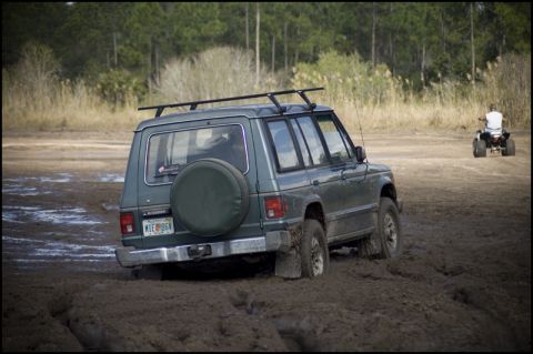1996 Ford F-150 - A green SUV navigates through a muddy terrain, while an ATV is seen in the distance.