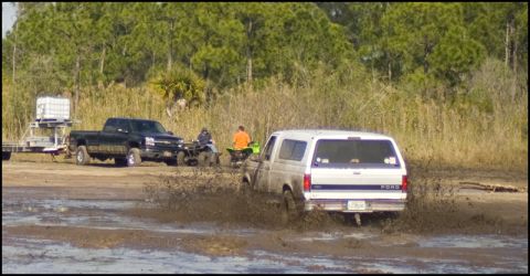 1996 Ford F-150 - A white Ford Bronco churns mud as it drives through a muddy area, with other vehicles and trees in the background.