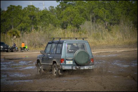 1996 Ford F-150 - A Mitsubishi Montero navigates a muddy off-road course, kicking up dirt as it goes. Other vehicles are visible in the background.