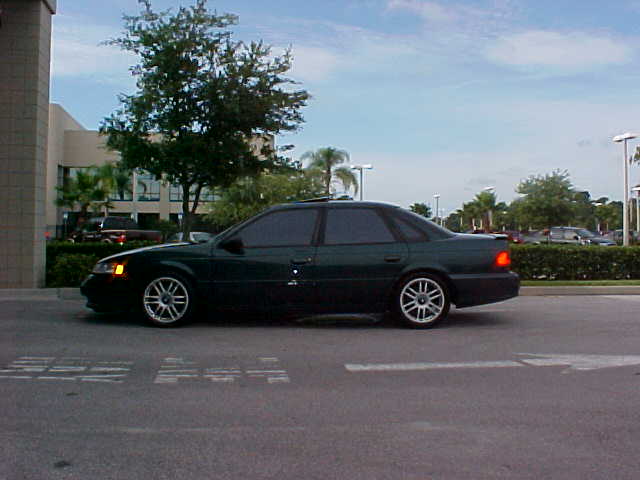 Dark green Ford Taurus sedan side profile with sunroof and multi-spoke wheels in a parking lot