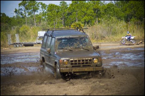 1996 Ford F-150 - An SUV navigates a muddy track, kicking up water, while a dirt bike rider passes by in the distance.