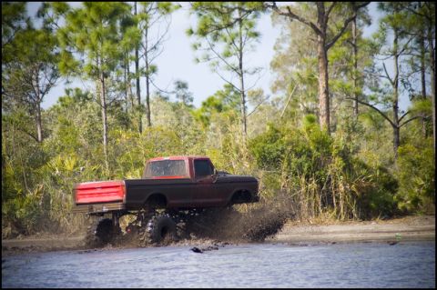 1996 Ford F-150 - Modified Ford F-Series pickup truck splashes through a muddy terrain with dense foliage in the background.