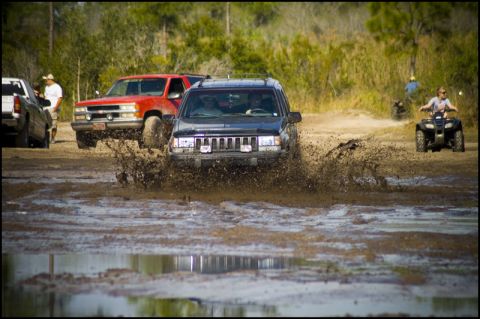 1996 Ford F-150 - Multiple vehicles navigate a muddy off-road course, kicking up large sprays of water and mud.