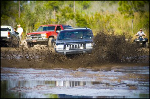 1996 Ford F-150 - Multiple off-road vehicles navigate a muddy track, kicking up large plumes of water and dirt.