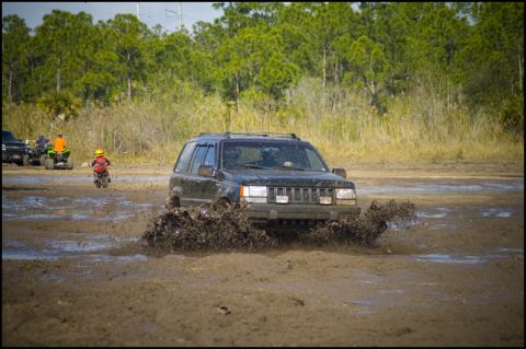 1996 Ford F-150 - A black Jeep Grand Cherokee navigates a muddy track, kicking up a large spray of dirt and water. Other off-road vehicles are visible in the…
