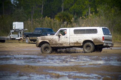 1996 Ford F-150 - A white pickup truck covered in mud navigates a muddy terrain, with another truck in the background.