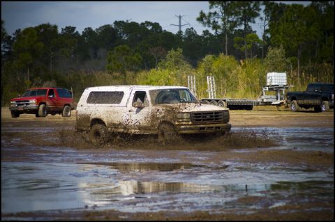 1996 Ford F-150 - A white SUV churns through mud, with other vehicles visible in the background.