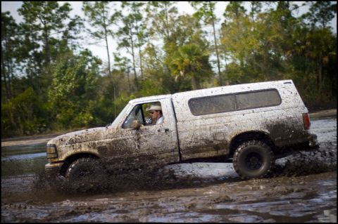 1996 Ford F-150 - A white pickup truck with a camper shell navigates a muddy off-road course, splashing water and mud.