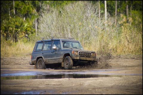 1996 Ford F-150 - A Mitsubishi Montero navigates a muddy off-road course, splashing water as it goes.