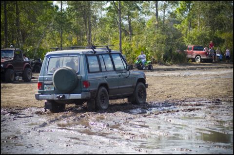 1996 Ford F-150 - Off-road vehicles navigate a muddy track with trees and spectators present.
